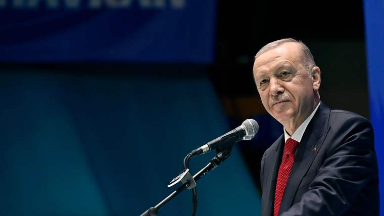 Kürsüde konuşan bir kişi, mikrofonun önünde duruyor; arka planda koyu mavi sahne bulunuyor.