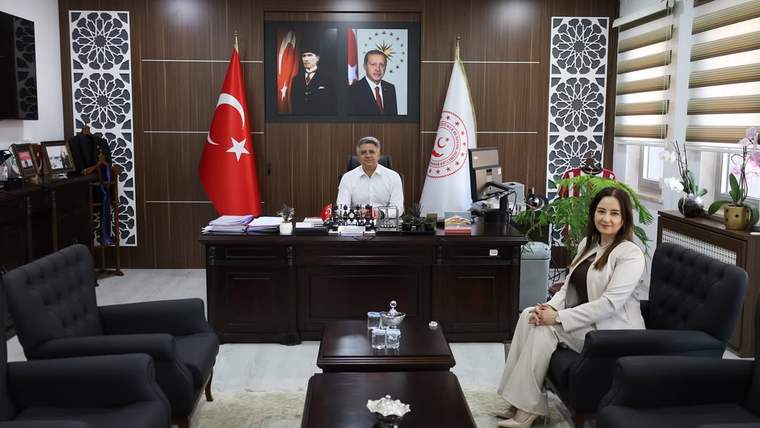 Türk bayrağı ve kurum flaması önündeki makam odasında masada oturan bir kişi ile koltuktaki kadın