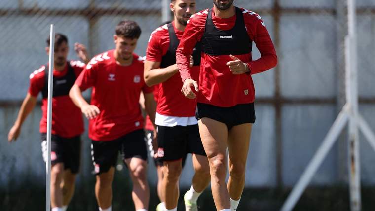 Antrenman sahasında kırmızı spor kıyafetler giyen bir grup erkek yan yana koşuyor.
