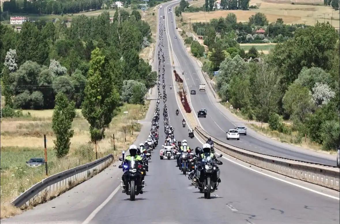 Çok sayıda motosikletli, bölünmüş kara yolunda ilerlerken çevrede ağaçlık ve tarım alanları görülüyor.