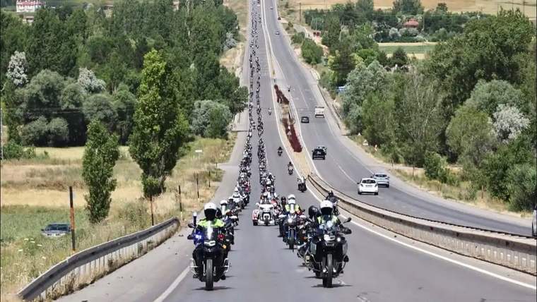 Çok sayıda motosikletli, bölünmüş kara yolunda ilerlerken çevrede ağaçlık ve tarım alanları görülüyor.