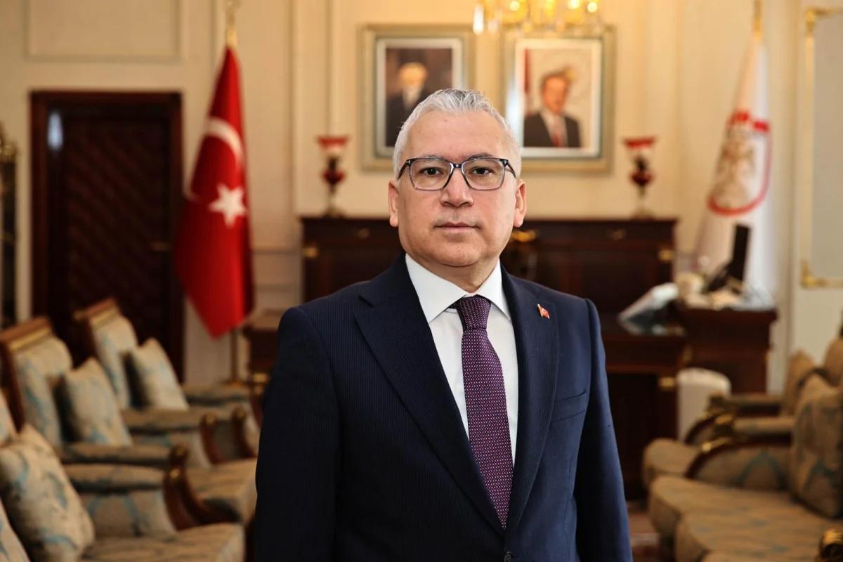 Takım elbiseli ve gözlüklü bir kişi, Türk bayrağı ile iki portre bulunan salonda duruyor.
