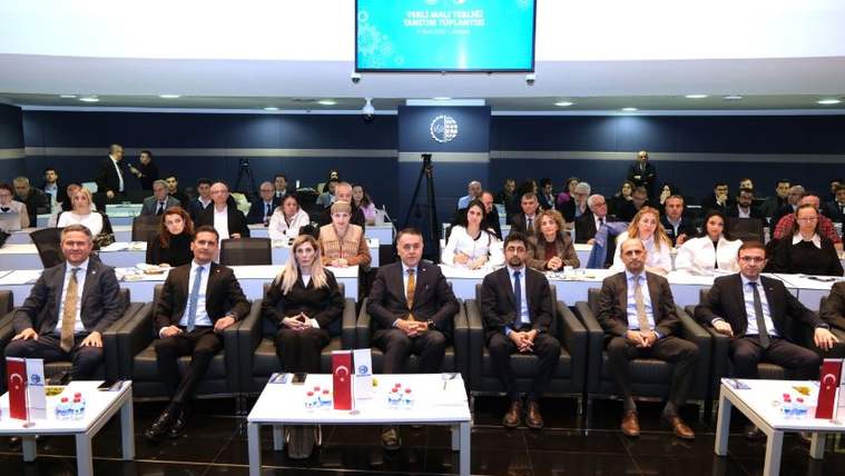 Bir toplantı salonunda, ön sırada oturan beş kişi ve arka planda katılımcılar yer alıyor.