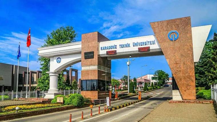 Karadeniz Teknik Üniversitesi yazılı giriş kapısı, bayrak direkleri ve kampüs yolu görülüyor.