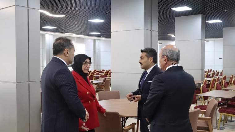 Geniş bir salonda dört kişi, masa ve sandalyelerin bulunduğu alanda ayakta sohbet ediyor.