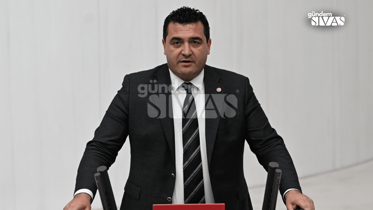 Kürsüde konuşan bir kişi, arka planda beyaz duvarlar bulunuyor.