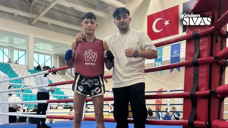 Boks ringinde iki kişi poz veriyor; genç olan koruyucu ekipman, diğeri beyaz tişört giyiyor.