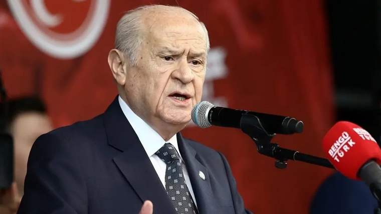 Takım elbiseli bir kişi, kırmızı arka plan önünde mikrofonlara konuşurken el hareketi yapıyor.