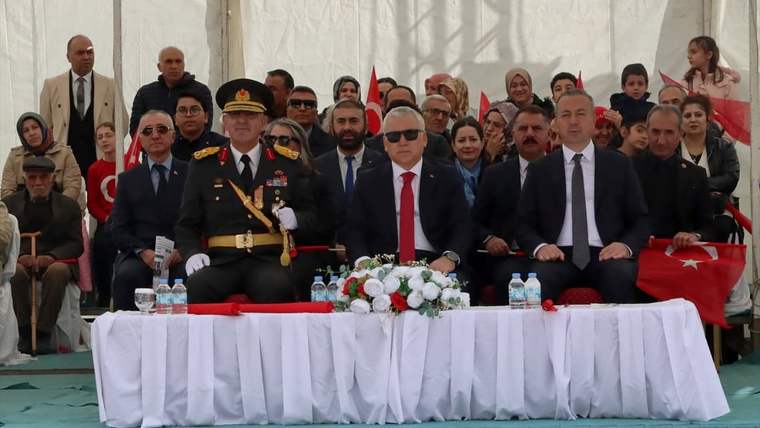 Ön sırada üniformalı bir kişi ve takım elbiseli kişiler, Türk bayrakları önünde masada oturuyor.
