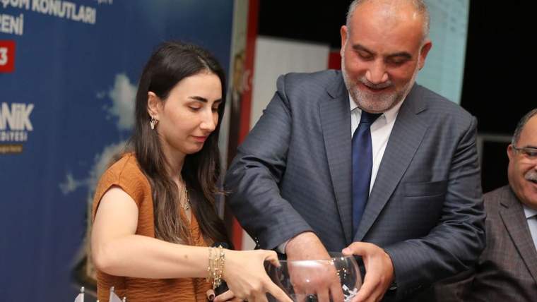 Bir kadın ve takım elbiseli bir erkek, içindeki kâğıtlarla birlikte şeffaf bir kura kabını tutuyor.