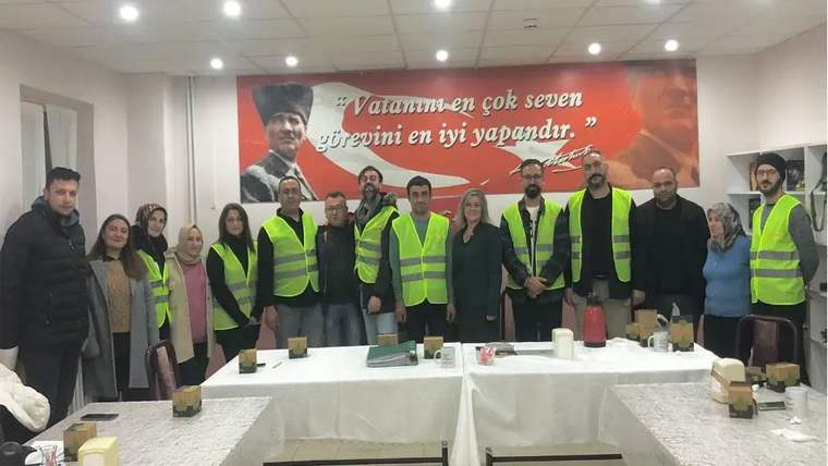 Toplantı salonunda, aralarında reflektörlü yelek giyenlerin bulunduğu bir grup, Atatürk posterli pankart önünde duruyor.