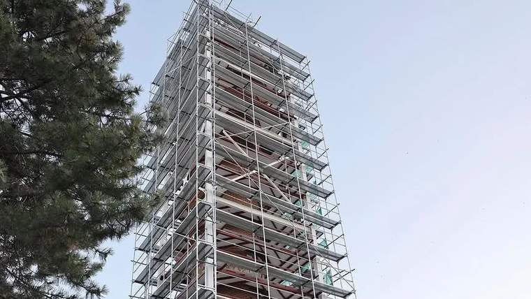 Çok katlı bir yapının çevresi metal iskelelerle sarılmış; sol tarafta bir çam ağacının dalları görülüyor.