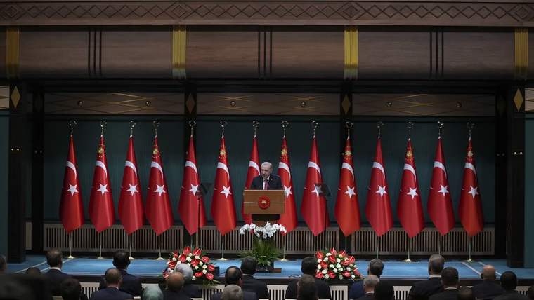 Bir kişi kürsüde konuşuyor, arkasında Türk bayrakları ve çiçekler var.