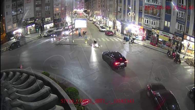 Gece aydınlatılmış şehir kavşağında araçlar ilerlerken yol ortasında bir kişi ve motosiklet görülüyor.