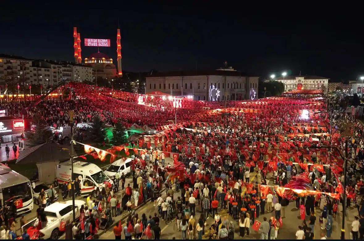 Bayraklarla süslenmiş kalabalık bir meydanda toplanmış insanlar, gece etkinliği.
