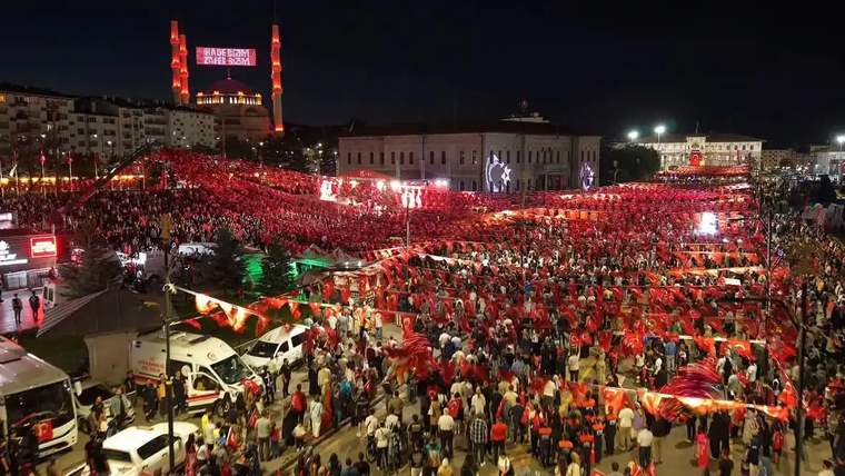 Bayraklarla süslenmiş kalabalık bir meydanda toplanmış insanlar, gece etkinliği.