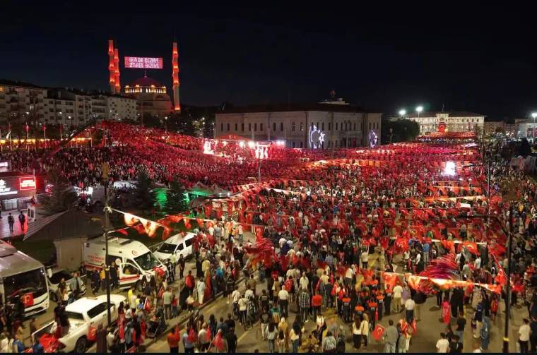 Bayraklarla süslenmiş kalabalık bir meydanda toplanmış insanlar, gece etkinliği.