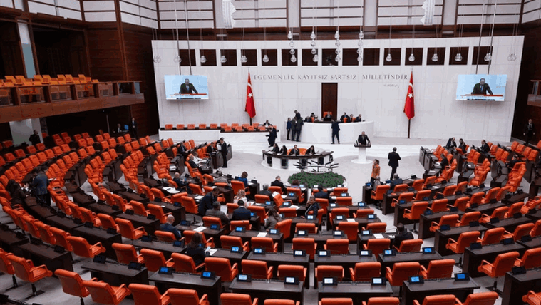 Genel kurul salonunda turuncu koltuklar, masalar, ekranlar, milletvekilleri ve kürsü görülüyor.