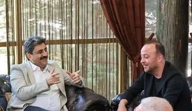 İç mekânda koltuklarda oturan iki erkek sohbet ederken biri elleriyle açıklama yapıyor.
