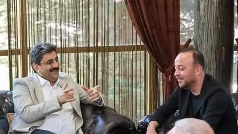 İç mekânda koltuklarda oturan iki erkek sohbet ederken biri elleriyle açıklama yapıyor.