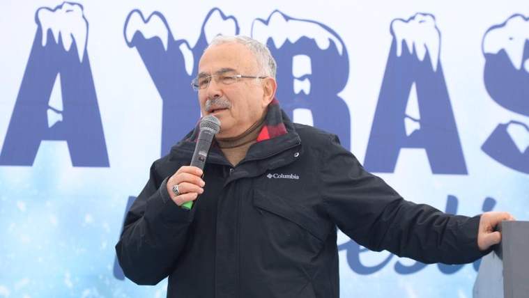 Mikrofonla konuşan gözlüklü bir kişi, kar desenli yazıların bulunduğu sahne önünde duruyor.