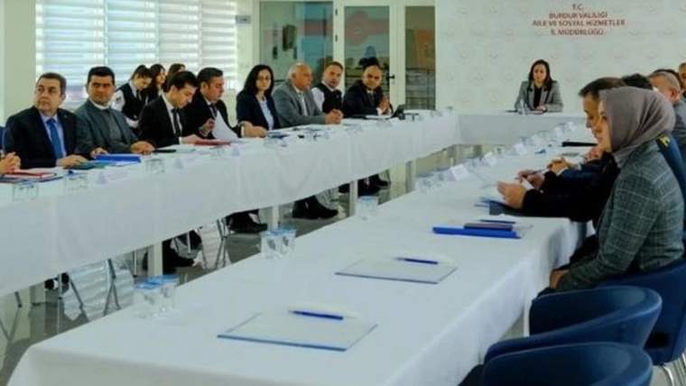 Uzun masaların çevresinde oturan kadın ve erkeklerden oluşan bir grup toplantı yapıyor; duvarda kurum tabelası bulunuyor.