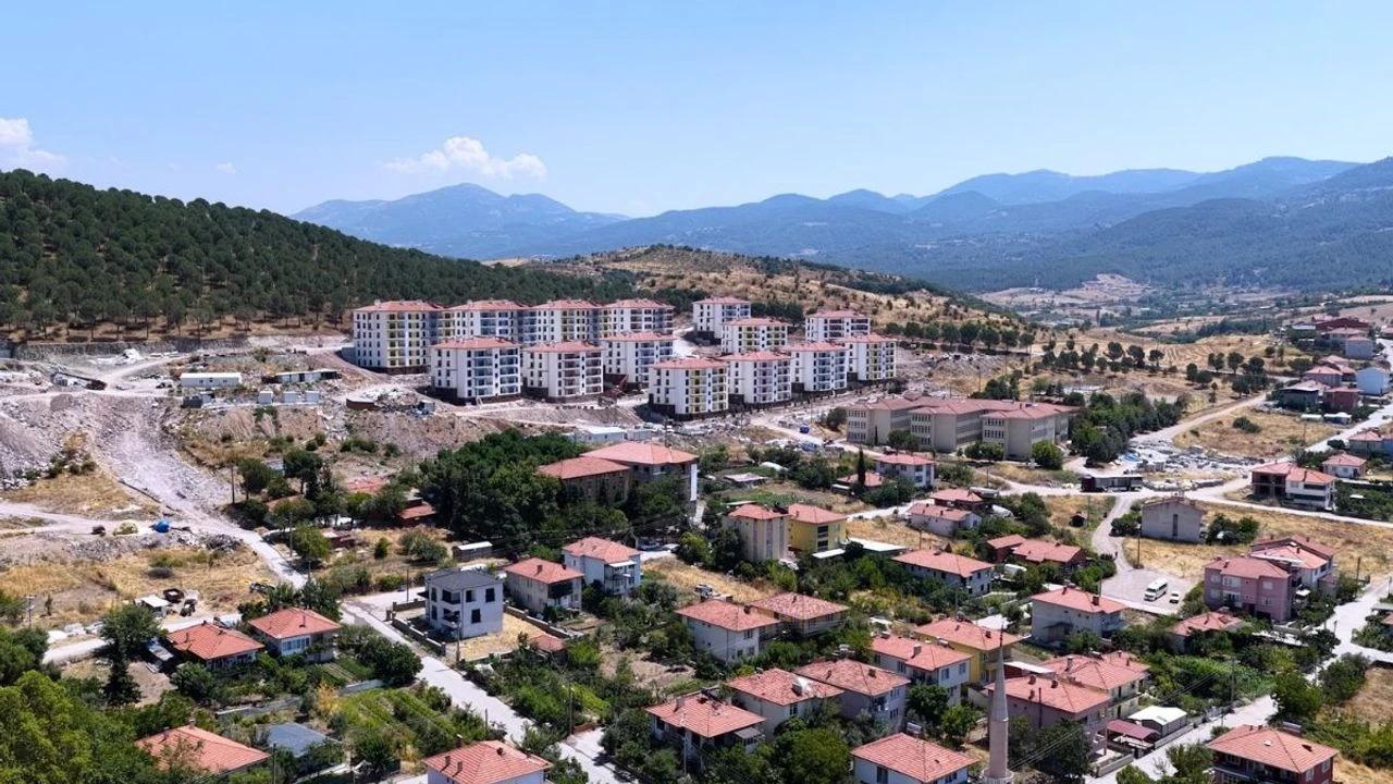 Dağlık arazinin önünde, çok katlı yeni binalar ve kırmızı çatılı evlerin bulunduğu yerleşim alanı