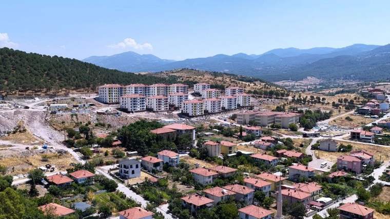 Dağlık arazinin önünde, çok katlı yeni binalar ve kırmızı çatılı evlerin bulunduğu yerleşim alanı