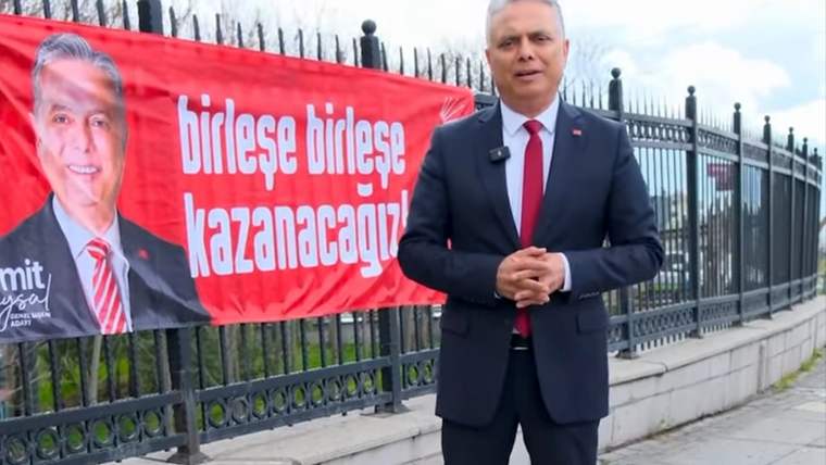 Takım elbiseli bir kişi, üzerinde “birleşe birleşe kazanacağız” yazan kırmızı pankartın önünde duruyor.