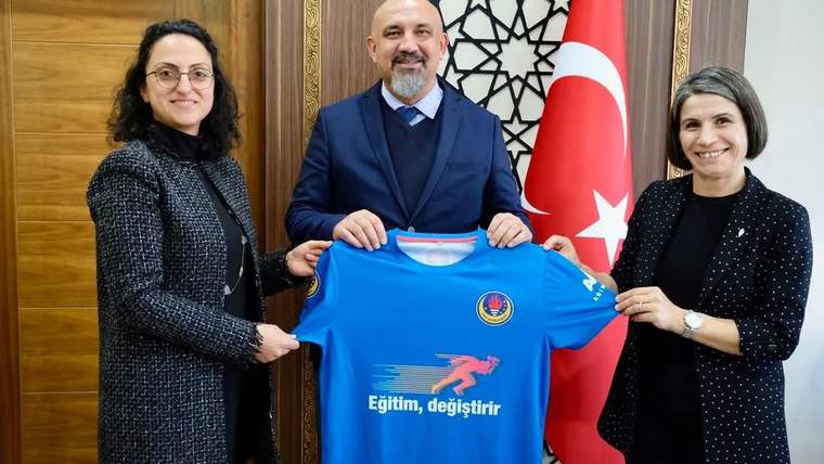 İç mekânda üç kişi, üzerinde Türk bayrağı amblemi ve “Eğitim, değiştirir” yazan mavi tişörtü tutuyor.