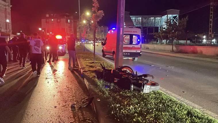 Yerde devrilmiş bir motosiklet, çevresinde ambulans ve kalabalık insanlar var.