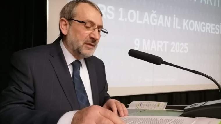 Takım elbiseli bir kişi, mikrofon önünde belgeleri incelerken arka planda kongre yazısı görülüyor.