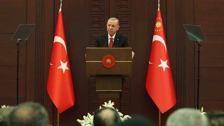 Kürsüde konuşan bir kişi, iki Türk bayrağı arasında duruyor; ön planda dinleyicilerin başları görülüyor.