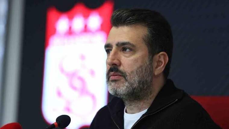 Siyah ceketli, sakallı bir kişi önündeki mikrofona bakarak oturuyor; arka planda bulanık bir amblem görülüyor.