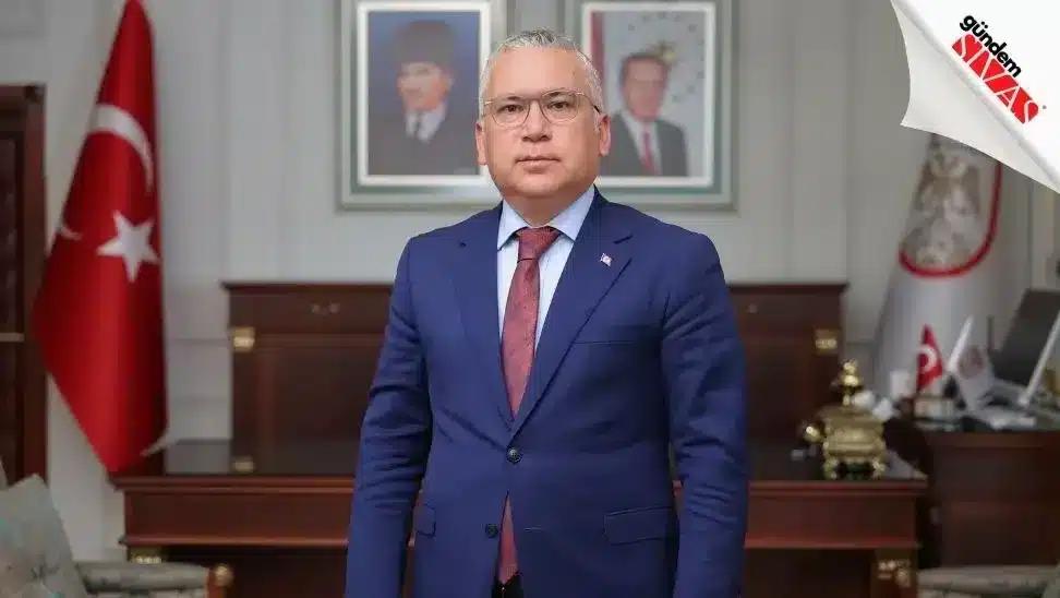 Mavi takım elbise giymiş bir kişi, arkasında Türk bayrağı ve çerçevelenmiş portreler ile poz veriyor.