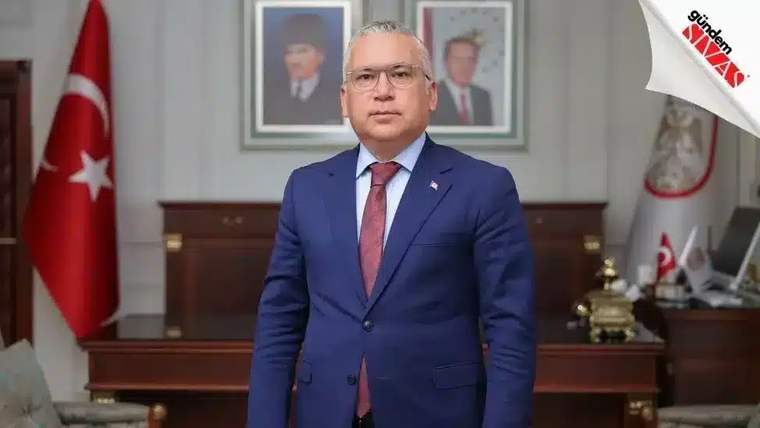 Mavi takım elbise giymiş bir kişi, arkasında Türk bayrağı ve çerçevelenmiş portreler ile poz veriyor.