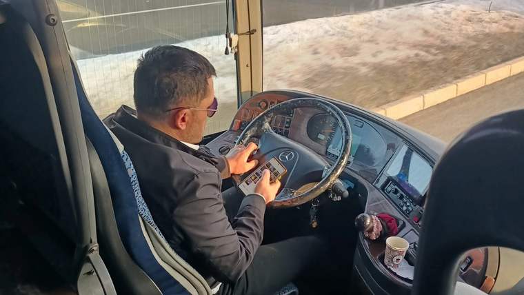 Direksiyon başında telefonla ilgilenen bir otobüs şoförü, otobüsün içi görünmekte.