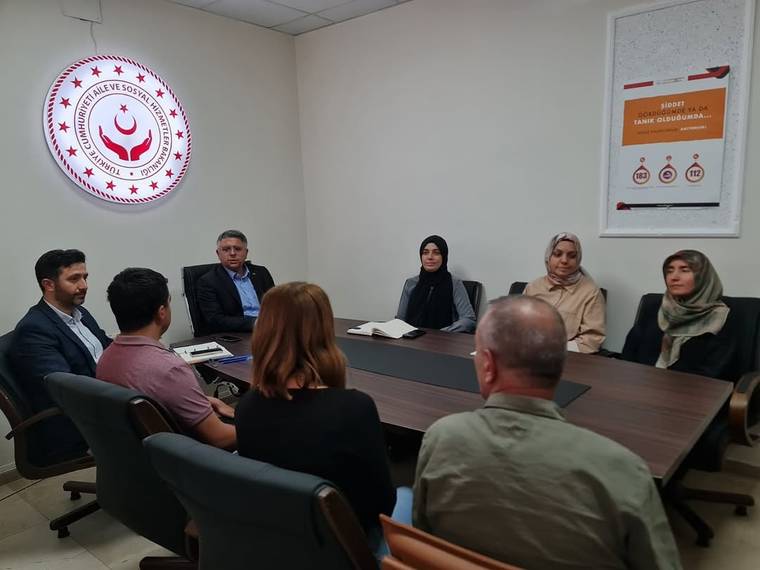 Toplantı masası çevresinde oturan altı kişi, duvarda kurum logosu ve bilgilendirici afiş bulunan odada görülüyor.