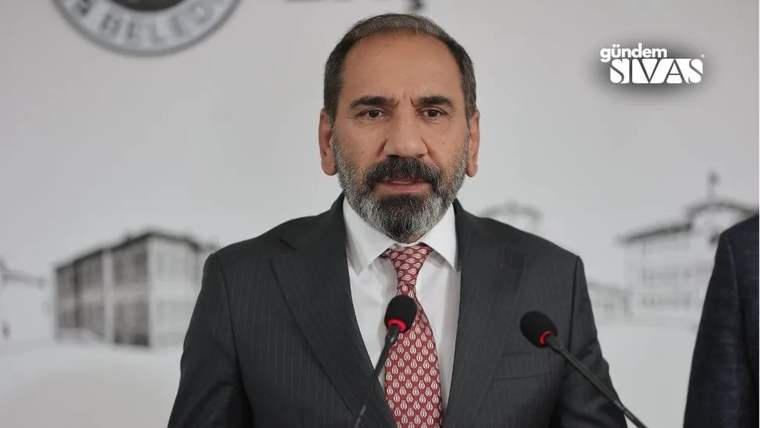 Kırmızı kravat takan, takım elbise giymiş bir kişi, mikrofonların önünde konuşuyor.