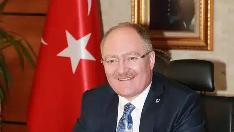 Masa başında oturan bir kişi, arkasında Türk bayrağı ve çerçeveli bir fotoğraf var.