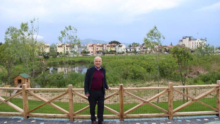 Bir kişi, ahşap bir korkuluk önünde duruyor; arka planda yeşil alan ve binalar görünmekte.