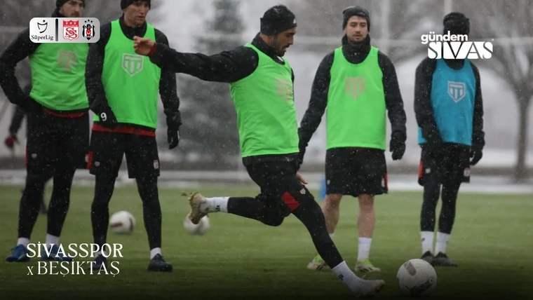 Yağmur altında futbol sahasında yeşil yelekli oyuncular antrenman yaparken biri topa vuruyor.