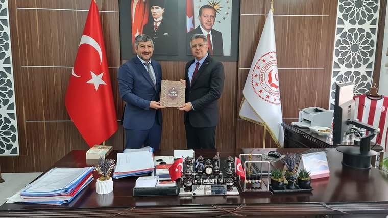 Makam odasında iki erkek, Türk bayrağı ve kurum flamaları önünde bir plaket tutarken görülüyor.