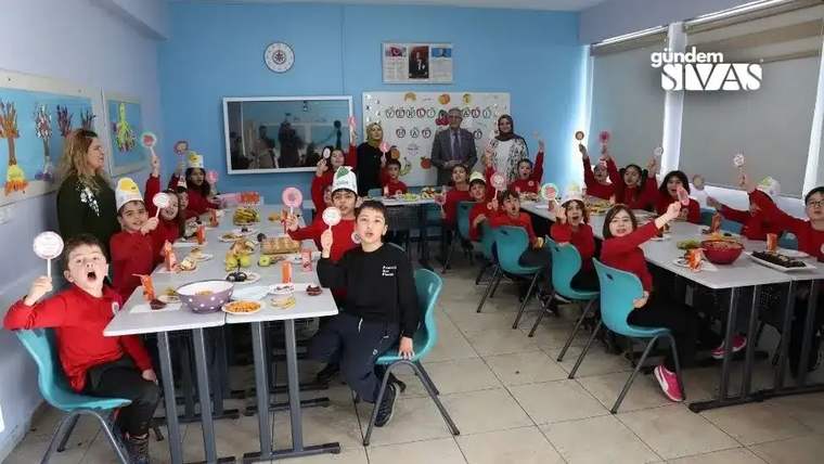 Sınıfta, kırmızı tişörtlü çocuklar, ellerinde yiyeceklerle gülümseyerek poz veriyor.
