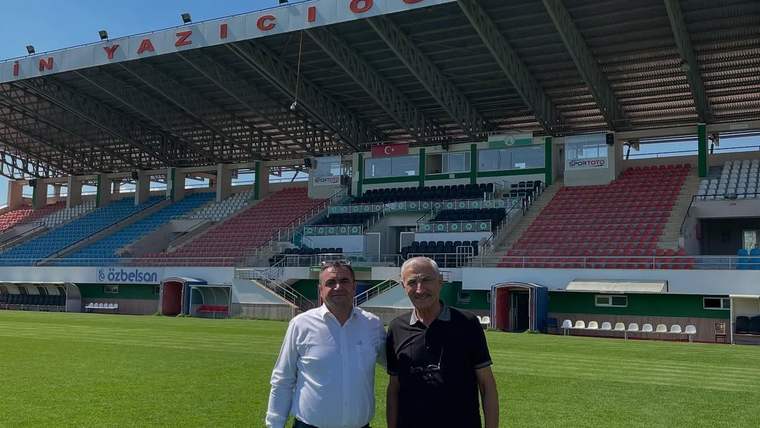 Tribünleri görünen futbol sahasının çimlerinde iki erkek yan yana duruyor; arkada koltuklar ve saha bölümleri yer alıyor.
