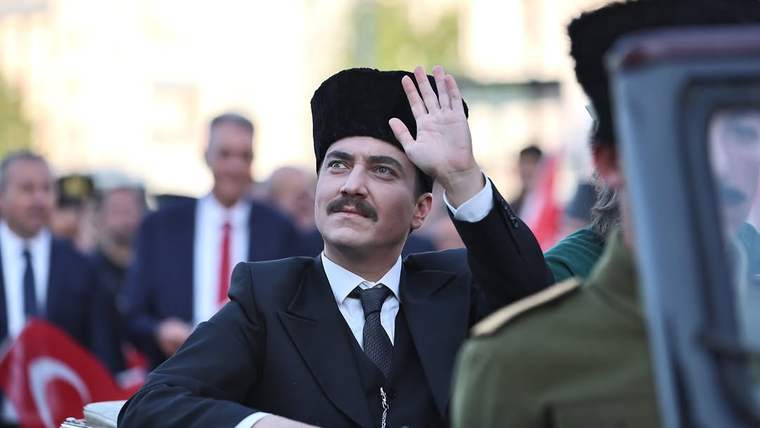 Siyah başlıklı ve takım elbiseli bir kişi, kalabalık arasında el sallarken görülüyor.
