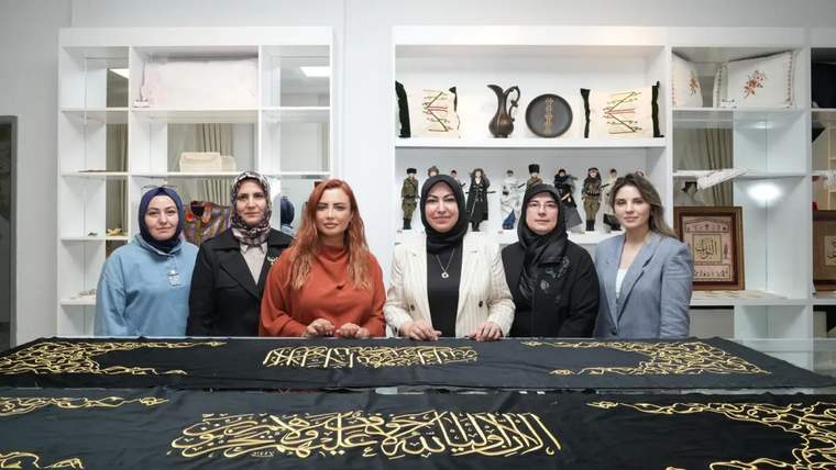 Altı kadın, siyah bir örtünün önünde duruyor; örtü üzerinde altın renkli yazılar var.