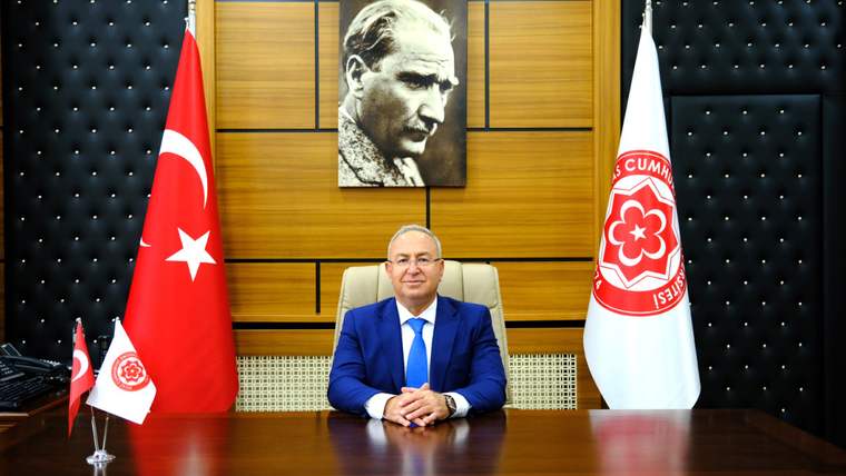 Ahşap panelli odada, Türk bayrağı ve amblemli beyaz bayrak önünde bir kişi masada oturuyor.