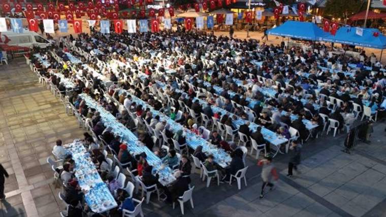 Uzun masalarda oturan kalabalık bir grup insan, iftar sofrasında yer alıyor.