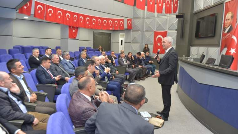 Bir kişi, mavi koltuklarda oturan kalabalığa konuşma yapıyor; arka planda Türk bayrakları asılı.
